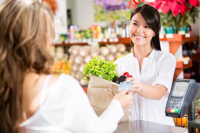 dependienta de comercio de proximidad con bolsa de papel con verduras, devuelve tarjeta bancaria a su clienta.